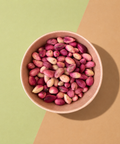 A top-down view of pink and cream pistachios in a light brown paper bowl, set against a diagonally split green and brown background.