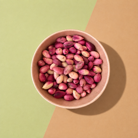 A top-down view of pink and cream pistachios in a light brown paper bowl, set against a diagonally split green and brown background.