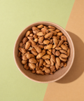 A top-down view of raw almonds piled in a small brown paper bowl, set against a split background of light green and tan paper.