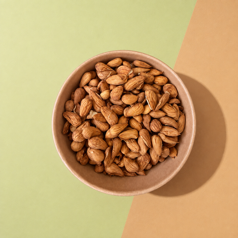 A top-down view of raw almonds piled in a small brown paper bowl, set against a split background of light green and tan paper.
